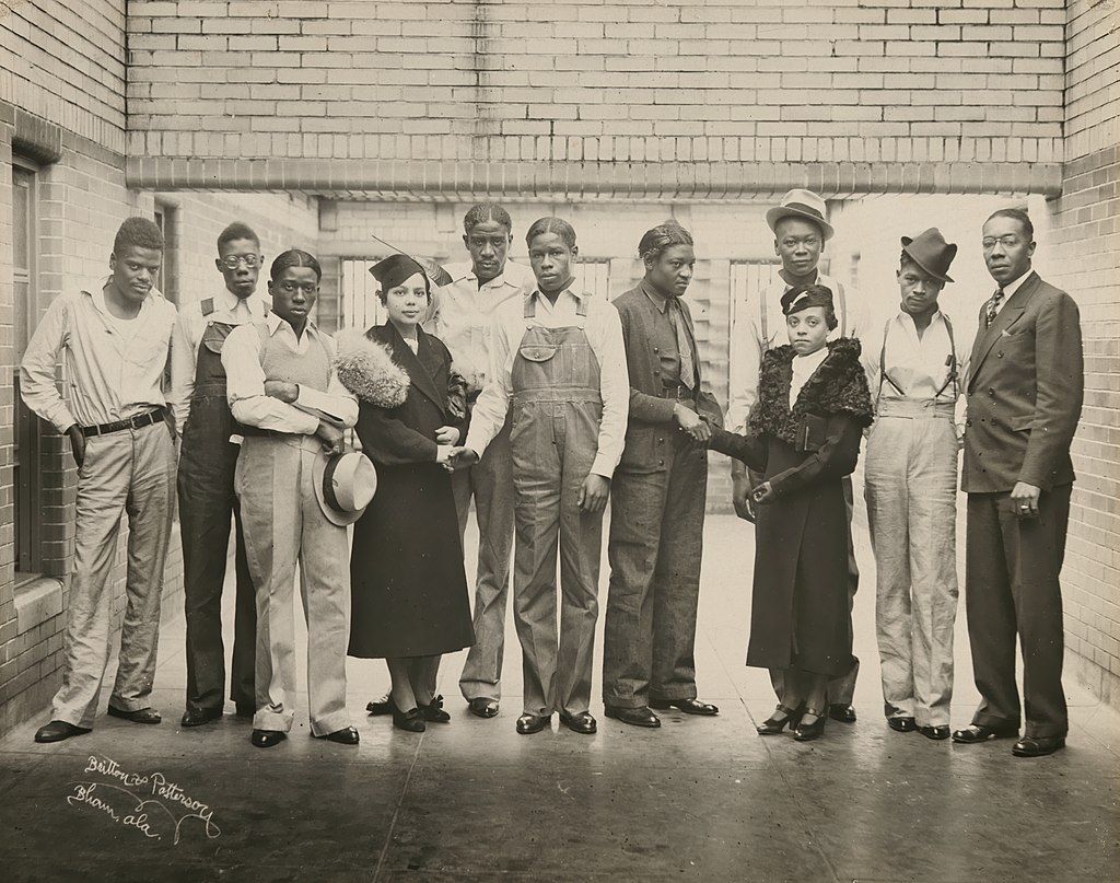 A black and white photo of 11 Black people in a hallway in the 1930s. They are the 9 Scottsboro Boys, Juanita Jackson Mitchell, and Laura Kellum. They are wearing or carrying tats and dressed variously in casual pants, overalls, and suits. 