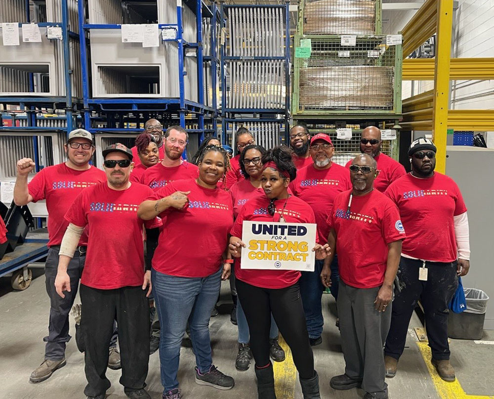 About a dozen workers in red shirts face the camera in an industrial space. The group is mostly Black, and a woman in front holds the sign saying, “United for a strong contract.
