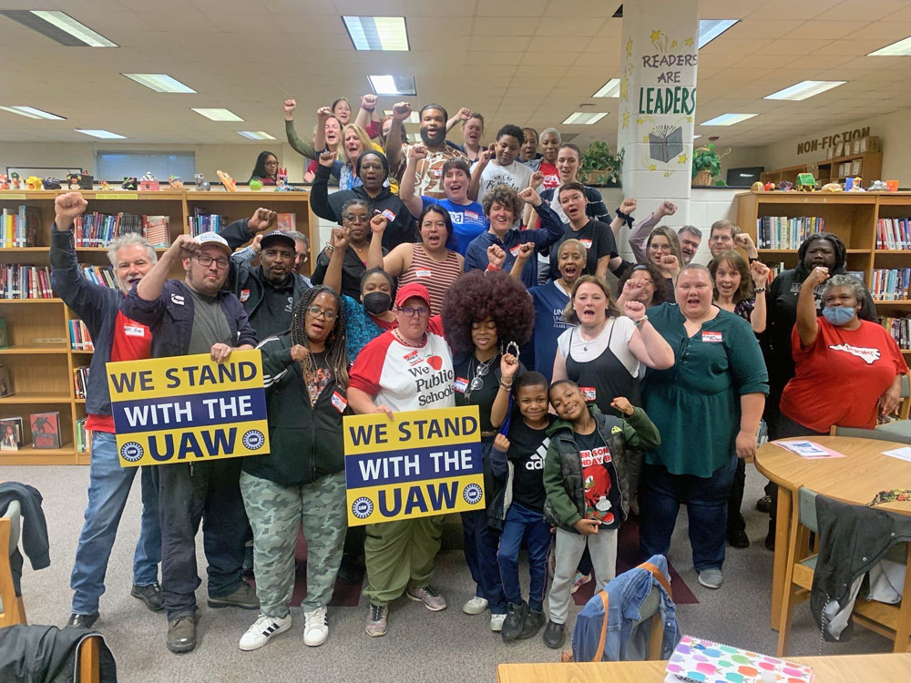 A crowd of some forty people—of mixed races and ages—in a library. Their fists are raised, and two hold signs that say, “We stand with the UAW.”