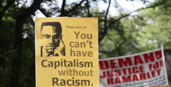 A yellow picket sign with black and grey printing reads Malcolm X: You can't have capitalism without racism. To the right, a white banner with red print reads Demand Justice for Ramarley.