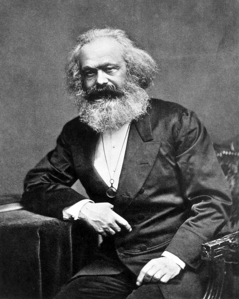 A black and white photograph of the philosopher and communist Karl Marx, an older white man with long gray hair and full beard. He is seated.