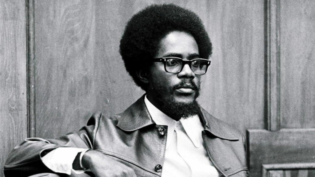 A black and white photograph of Guyanese socialist Walter Rodney, a black man with a mid-lenth Afro and black glasses. He is wearing a leather coat and is seated.