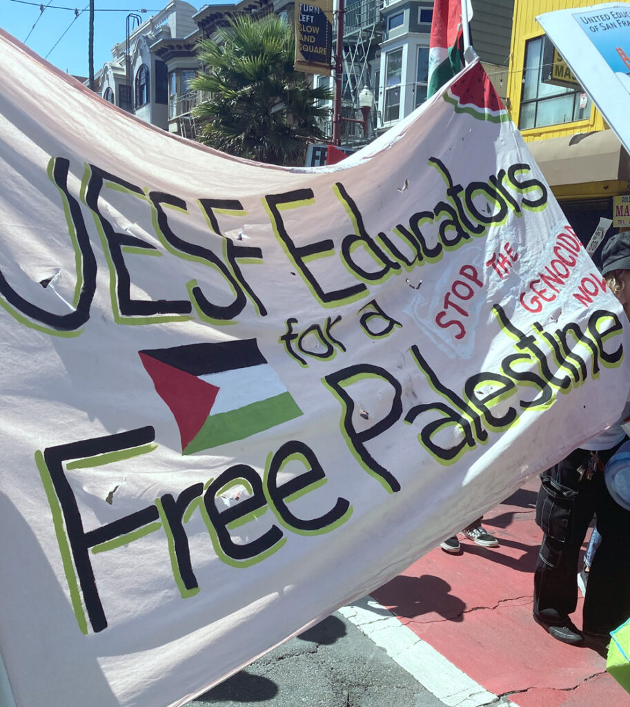Hand-painted banner on a brightly-lit street saying, “UESF Educators for a Free Palestine: Stop the genocide now.” Banner decorated with a Palestinian flag near the middle and a watermelon wedge in the upper right corner.